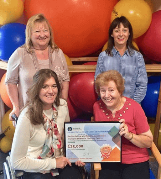 Four ldies, two standing and two seated in front of colourful gym balls, holding a framed cheque for £25, 000